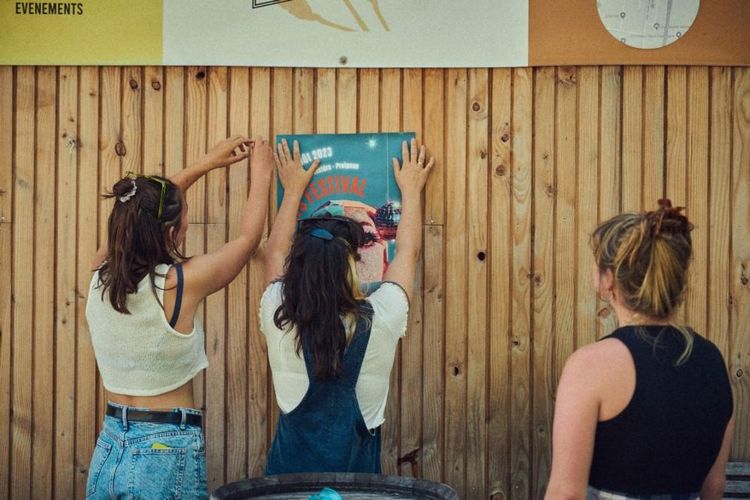 Trois filles du collectif de dos, en train de coller les affiches du festival sur une palissade en bois